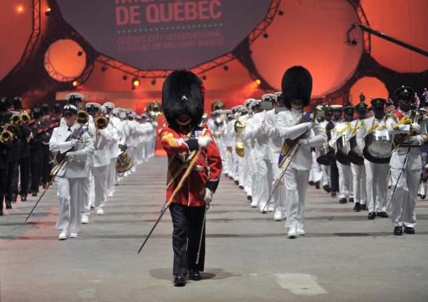 Eine Gruppe uniformierter Personen marschiert auf einer Straße, einige spielen Musikinstrumente, mit einem beleuchteten Schild im Hintergrund während der Eröffnungszeremonie des Montreal International Festival of Military Bands.