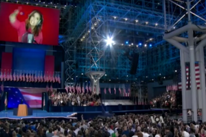 Eine große Menschenmenge sitzt vor einer Bühne mit einem Podium, einem Mikrofon, Flaggen und einem Bildschirm mit dem Bild einer Frau, der auf eine Präsidenten-Debatte wartet.