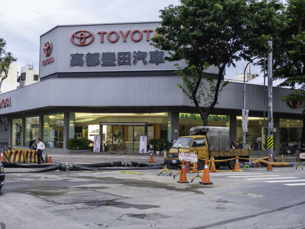 Eine Toyota-Händlerwerkstatt in Tokio, Japan, mit Fahrzeugen, Verkehrskegeln, Rohren, Menschen, Bäumen, Pfählen, Platten und Gebäuden im Vordergrund und einem bewölkten Himmel im Hintergrund.