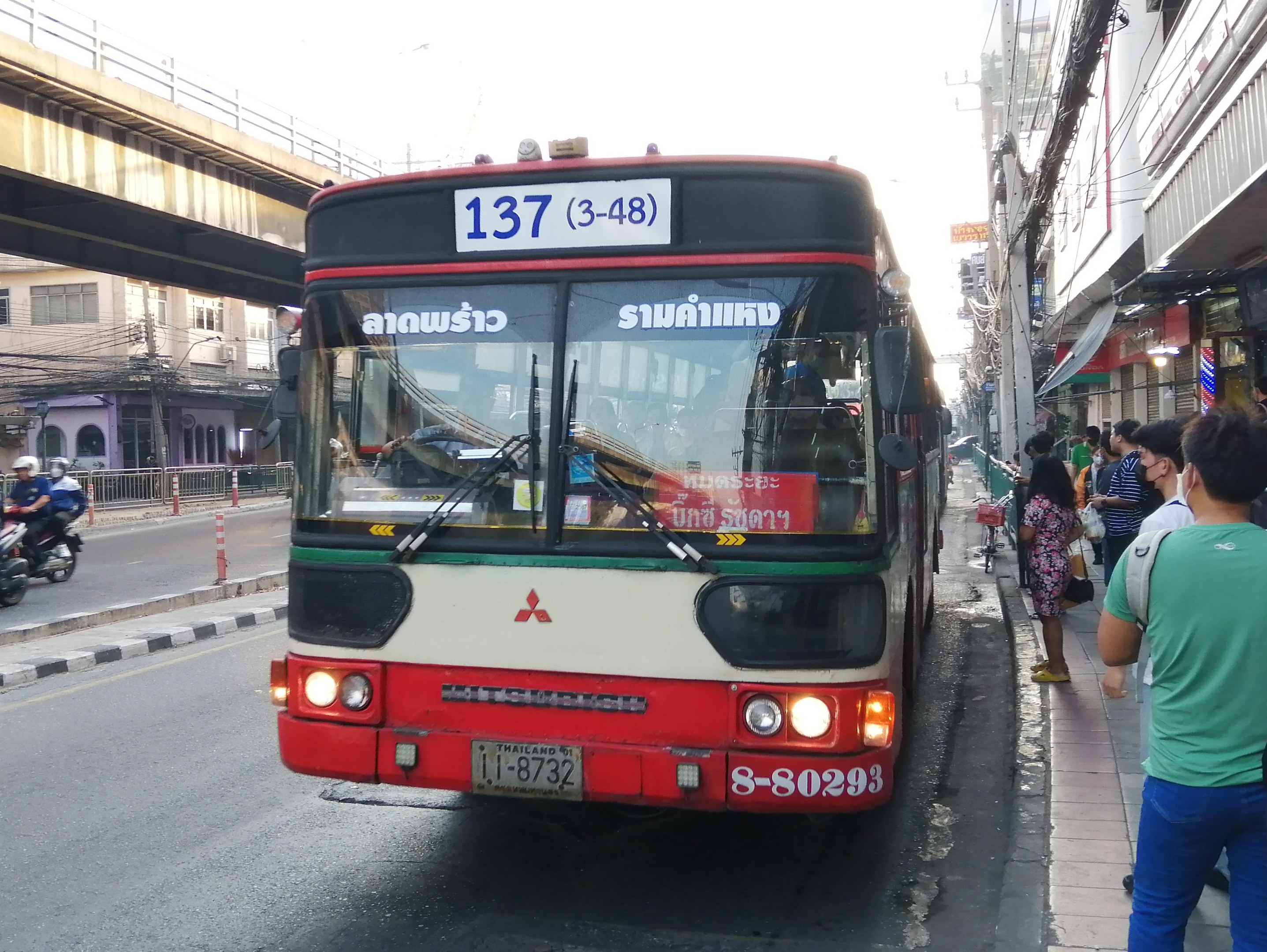 Ein roter und weißer Bus fährt eine Straße mit hohen Gebäuden entlang, Fußgänger auf dem Gehweg, eine Brücke, Gebäude, Strommasten mit Drähten und einen klaren blauen Himmel.