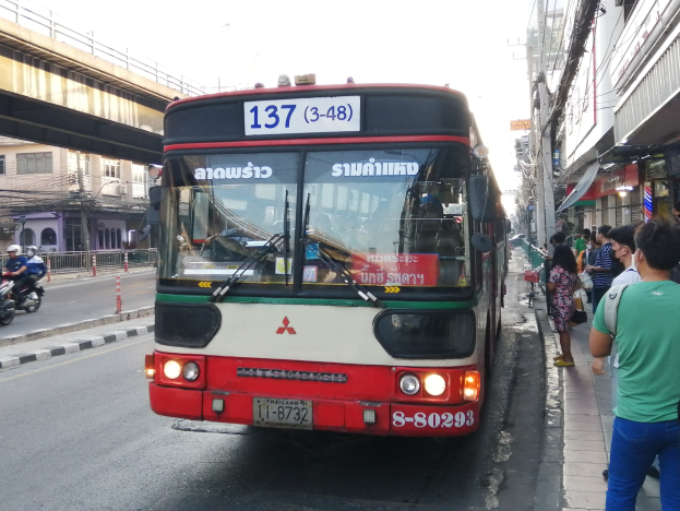 Ein roter und weißer Bus fährt eine Straße mit hohen Gebäuden entlang, Fußgänger auf dem Gehweg, eine Brücke, Gebäude, Strommasten mit Drähten und einen klaren blauen Himmel.