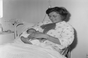 Eine Frau in einem Krankenhausbett hält ein Neugeborenes, umgeben von einem Kissen und einer Decke, mit einem Tisch links und Rohren im Hintergrund.