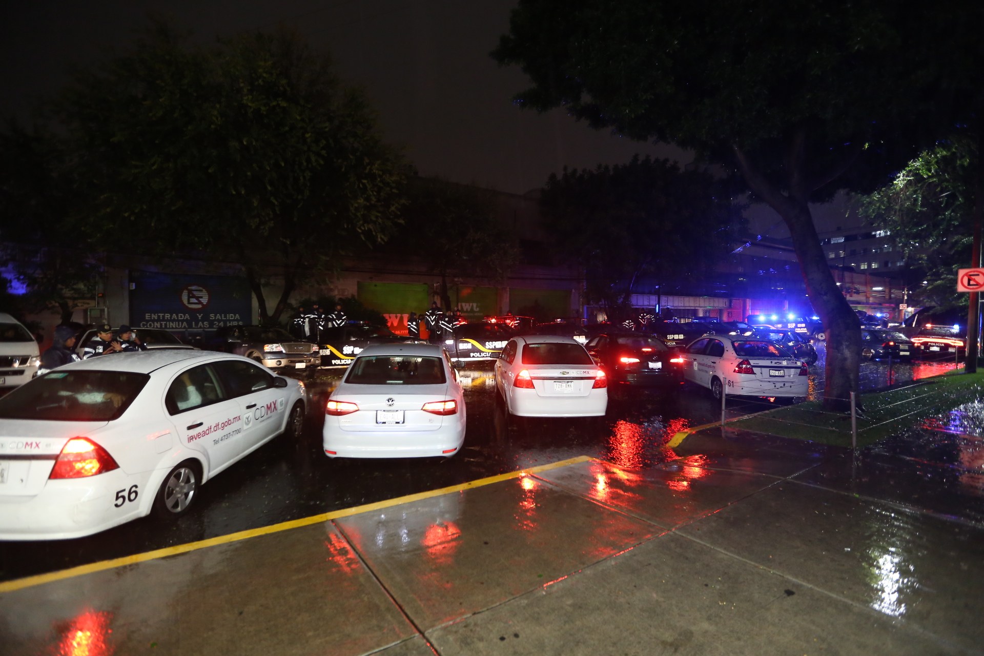 Polizeiautos auf einem Parkplatz bei Nacht mit Gebäuden, Bäumen, einem Schild und einer Fahne im Blickfeld unter einem bewölkten Himmel.