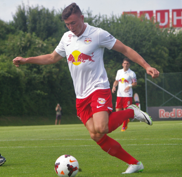 Ein Fussballer in einem Fussballtrikot schießt einen Ball auf einem grünen Feld mit Bäumen und einem klaren blauen Himmel im Hintergrund, wobei ein Schild mit der Aufschrift "RB Leipzig" sichtbar ist.