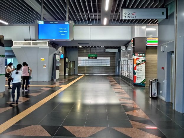 Menschen mit einem Koffer durch einen Bahnhof mit einer Bahre auf der rechten Seite, Schilder mit Text, eine Uhr an der Wand und Deckenbeleuchtung.