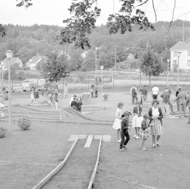 Schwarz-weißes Bild von Menschen, die auf einem Parkgelände mit Grünflächen, Laternen, geparkten Fahrzeugen und Gebäuden im Hintergrund Minigolf spielen.
