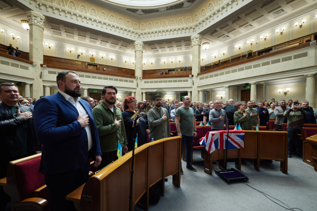Eine Gruppe von Liberalen, die im House of Commons steht, mit einer Menge, Stühlen, Tischen, Flaggen, einem Mikrofonständer, einem Gerät auf dem Boden, Säulen, Wandlampen und Deckenlampen im Hintergrund.