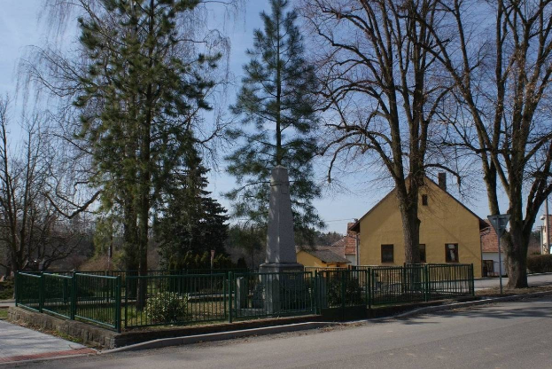 Eine Straße mit einem Holocaust-Denkmal in der Mitte, umgeben von einem Metallzaun, umgeben von Gras, Bäumen, Häusern, einem Straßenpfahl und einem bewölkten Himmel.