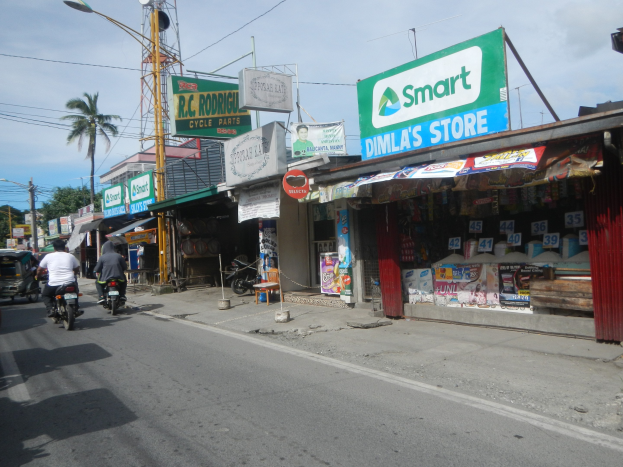 Eine belebte Straße mit Motorrädern, Autos, Gebäuden, Straßenschildern, Strommasten, Drähten, Laternen, Bäumen und einem bewölkten Himmel.