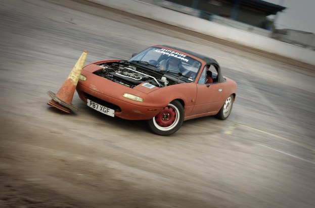 Eine Person in einem roten Auto auf einer Rennstrecke mit einem Verkehrskegel vorneweg, der Hintergrund unscharf, was auf hohe Geschwindigkeit hinweist.