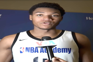 Ein Mann in einem weißen Tanktop hält ein Mikrofon vor ein Banner mit der Aufschrift "NBA All-Star Game 2019" und spricht mit der Medien.