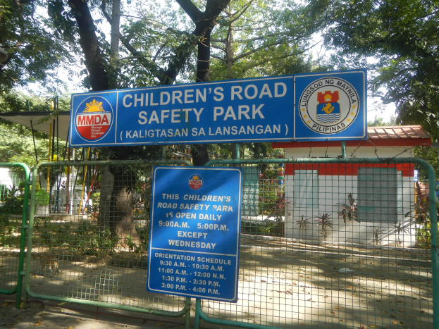 Ein von einem Metallzaun umgebener Kinderverkehrssicherheitspark mit zwei blauen Informationsschildern, einem kleinen Haus mit Dach, einigen Pflanzen, einer Baumgruppe und einem bewölkten Himmel.