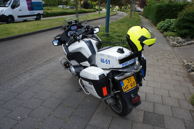 Ein Polizei-Motorrad am Straßenrand geparkt mit einem Helm darauf, umgeben von Bäumen, Pfählen, Gebäuden, Pflanzen, Gras, Steinen und anderen Fahrzeugen.