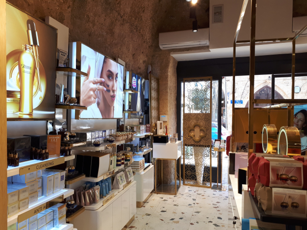 Ein gut beleuchteter Kosmetikgeschäftsinnenraum mit Regalen, auf denen verschiedene Schönheitsprodukte stehen, an der Wand montierte Bildschirme, Deckenleuchten, eine Klimaanlage und Glastüren, die eine Person, ein Auto und ein Gebäude draußen zeigen.