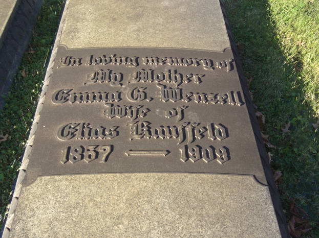 Ein Grabstein auf einem Friedhof mit der Inschrift "Die liebevolle Erinnerung an meine Mutter", umgeben von Gras und trockenen Blättern.