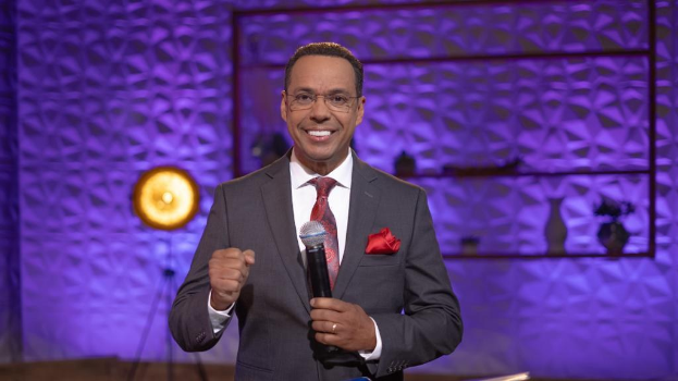 Stephen Colbert steht auf einer Buhne und halt einen Mikrofon in der Hand, tragt einen Anzug, Krawatte und Brille, mit einem Scheinwerfer und Stativ hinter ihm.