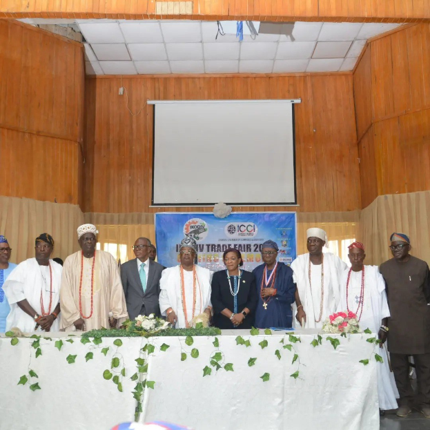 Gruppe von Menschen, die um einen weißgedeckten Tisch mit Blumen und Blättern stehen, mit einem Banner mit der Aufschrift "ICCI International Trade Fair 2019" im Hintergrund, neben einem Projektionsschirm und Deckenbeleuchtung.
