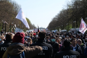 Eine große Gruppe von Menschen steht vor einer Menge von Polizeibeamten, einige tragen Mützen und Masken, während einer Demonstration in Berlin, Deutschland, mit Transparenten, Fahnen, Laternenmasten, Bäumen, Fahrzeugen, einem Gebäude und dem Himmel im Hintergrund.