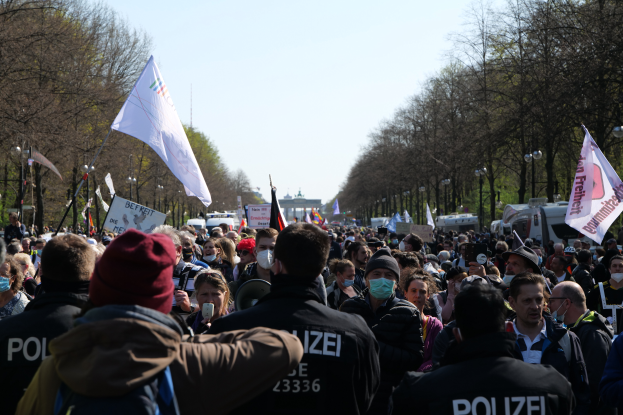 Eine große Gruppe von Menschen steht vor einer Menge von Polizeibeamten, einige tragen Mützen und Masken, während einer Demonstration in Berlin, Deutschland, mit Transparenten, Fahnen, Laternenmasten, Bäumen, Fahrzeugen, einem Gebäude und dem Himmel im Hintergrund.