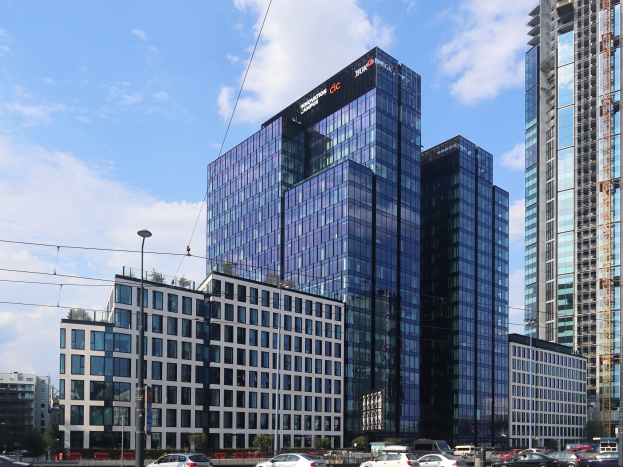 Zentrale der Deutschen Bank in Frankfurt, Deutschland, ein großes Gebäude mit vielen Fenstern, umgeben von städtischer Infrastruktur einschließlich Straßenlaternen, Fahrzeugen, Grünflächen und einem bewölkten Himmel.