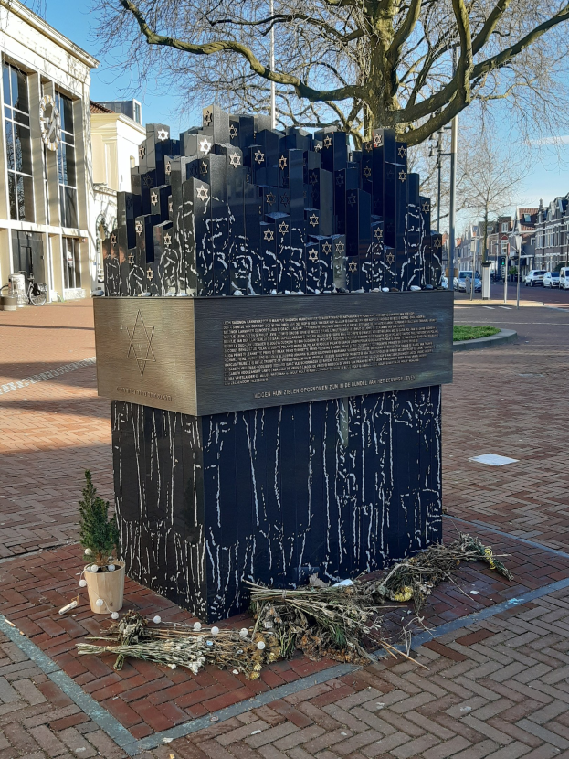 Denkmal für Holocaust-Opfer in einem Stadtplatz mit einem Baum davor, Topfpflanze, verstreuten trockenen Blättern und umgebenden Gebäuden, Fahrzeugen, Masten und Bäumen.