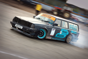 Ein Auto fährt auf einer Rennstrecke, wobei Rauch ausgestoßen wird, umgeben von anderen Fahrzeugen und Menschen, alles unscharf aufgrund der hohen Geschwindigkeit.