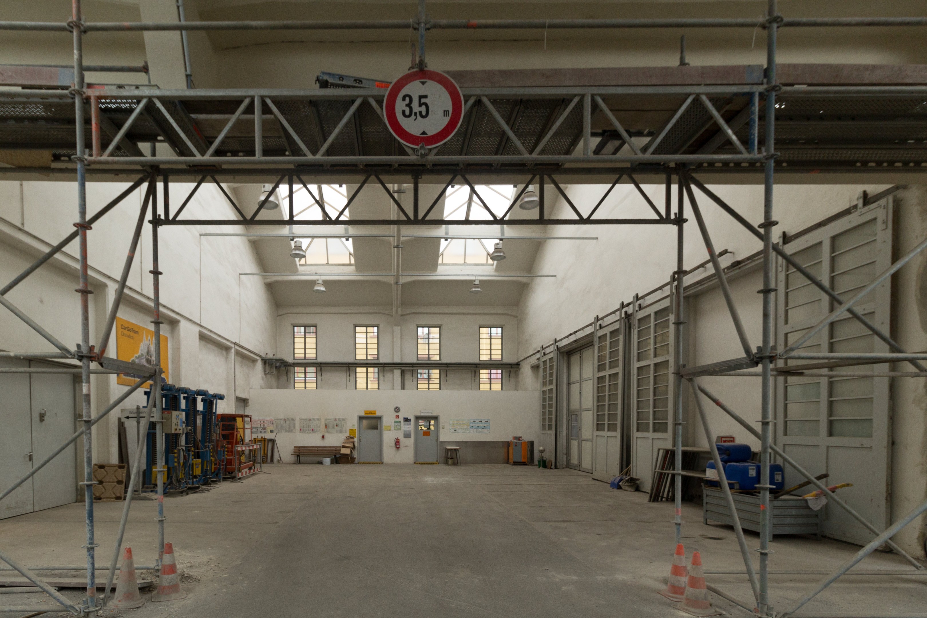 Lagerhalle mit Gerüst, Verkehrskegeln, Metallstangen, einem Schild, Maschinen, einer Tür, Fenstern, einer Wand, Deckenleuchten und einem Dach.