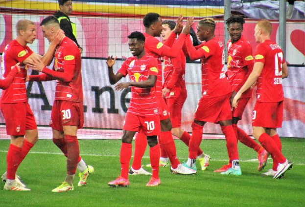 Eine Gruppe von Fußballspielern feiert ein Tor auf einem Rasenfeld, mit einem Netz, Bannern mit Text, Pfosten und anderen Gegenständen im Hintergrund.