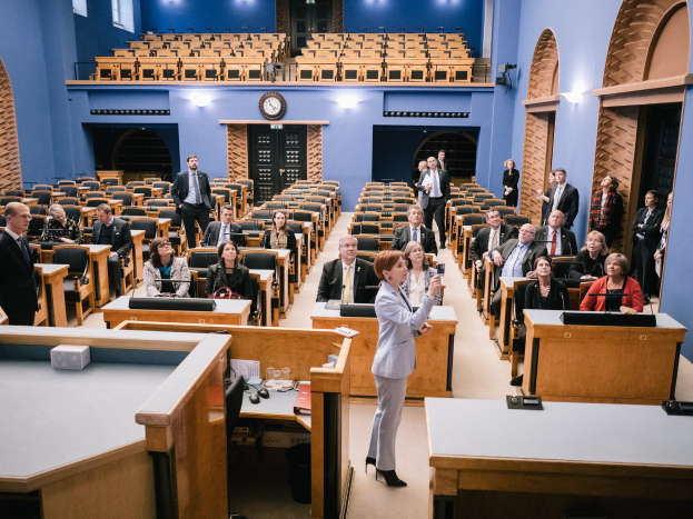 Frau hält Rede im schottischen Parlament vor einem Publikum, das auf Stühlen sitzt und steht, mit Mikrofonen, Gläsern, Papieren und anderen Gegenständen auf den Tischen, einer Uhr an der Wand und Deckenleuchten im Hintergrund.