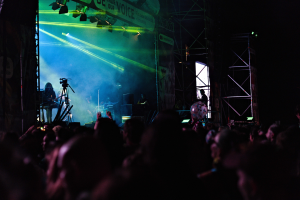 Eine Menge schaut Performern auf einer hell erleuchteten Bühne mit Musikinstrumenten, Mikrofonen und einem Banner bei einem Konzert zu.