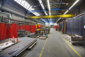 Großes Lager mit Holzpaletten, Metallobjekten, Verkehrskegeln und Maschinenfahrzeug, mit roten Vorhängen, Rohren, Deckenleuchten und einer Vorhangwand.