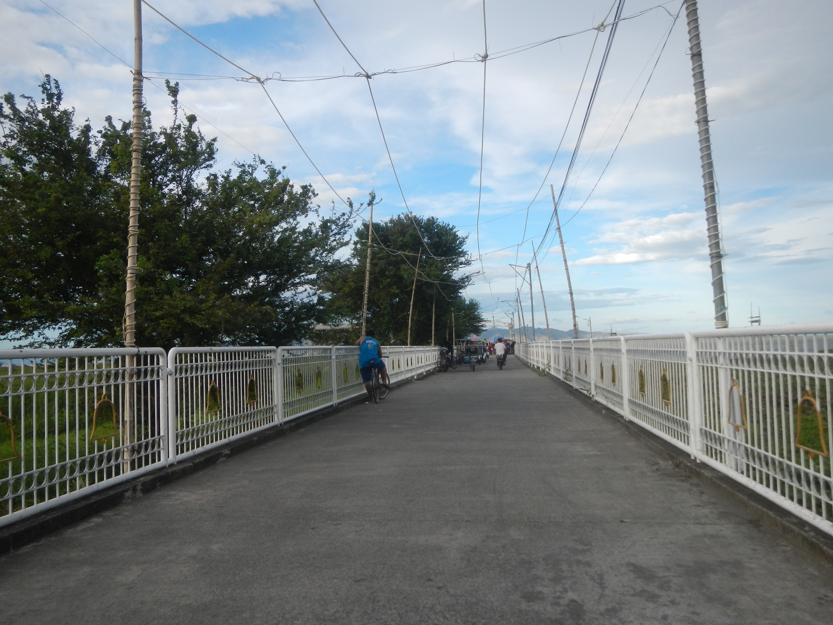 Eine Fußgängerbrücke mit Radfahrern, Geländern, Strommasten, Bäumen und einem bewölkten Himmel im Hintergrund.