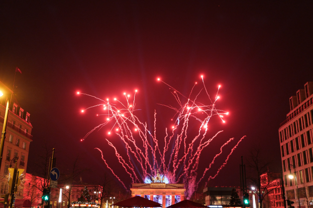Eine nächtliche Straßenansicht in Berlin an Silvester mit Gebäuden, Bäumen, Laternen, Verkehrszeichen, Schildern, Zelten, Menschen und einem beeindruckenden Feuerwerk am Himmel.