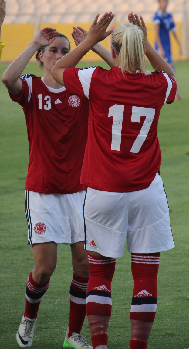 Zwei Frauen in roten T-Shirts und weißen Shorts stehen auf einem Fußballfeld und klatschen in die Hände, im Hintergrund ist eine Person in einem blauen Kleid, eine gelbe Tafel und einige Stühle zu sehen.