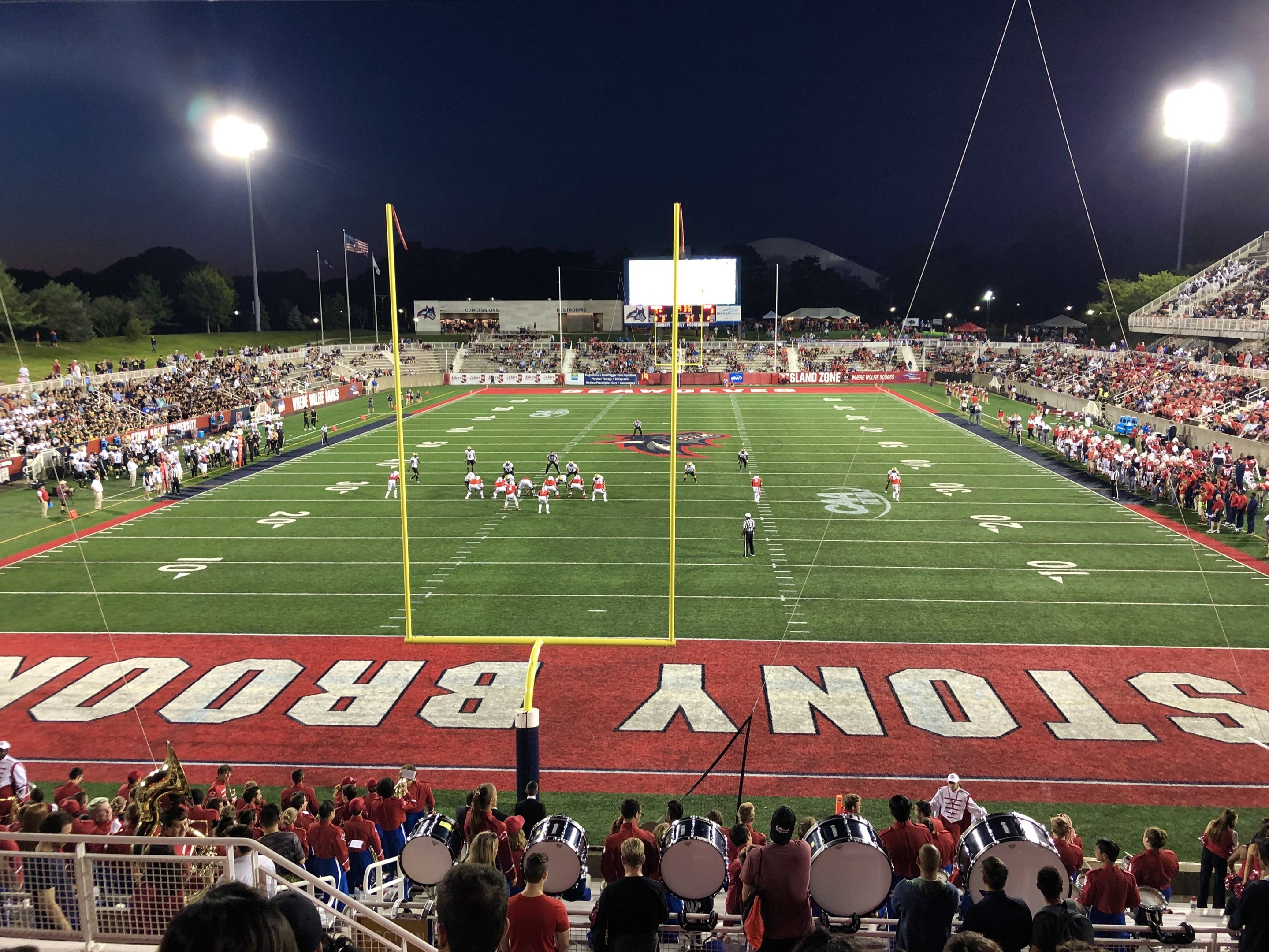 Ein nächtliches Fußballspiel in einem Stadion mit Spielern auf dem Feld, Zuschauern auf Stühlen und Musikern mit Instrumenten, umgeben von einem Metallzaun mit Laternen, Fahnen, einem Display, Bäumen, Hügeln und einem sternenklaren Himmel.