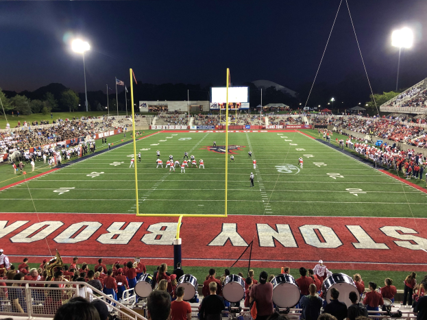 Ein nächtliches Fußballspiel in einem Stadion mit Spielern auf dem Feld, Zuschauern auf Stühlen und Musikern mit Instrumenten, umgeben von einem Metallzaun mit Laternen, Fahnen, einem Display, Bäumen, Hügeln und einem sternenklaren Himmel.