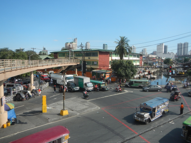 Eine belebte Stadtstraße mit Autos, Bussen, Motorrädern, einer Brücke mit Geländern und Pfeilern, Laternen, Strommasten mit Drähten, Gebäuden mit Fenstern, Bäumen und einem bewölkten Himmel.