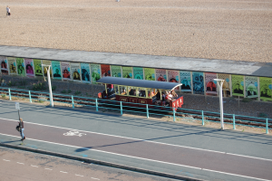 Ein kleiner Zug mit Passagieren fährt auf Schienen neben einem Strand, mit einer Person, die auf einem Fußweg zu links geht und einer Wand mit Plakaten im Hintergrund.