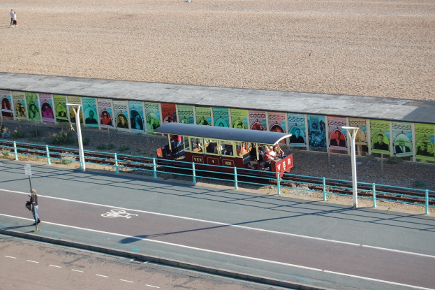 Ein kleiner Zug mit Passagieren fährt auf Schienen neben einem Strand, mit einer Person, die auf einem Fußweg zu links geht und einer Wand mit Plakaten im Hintergrund.