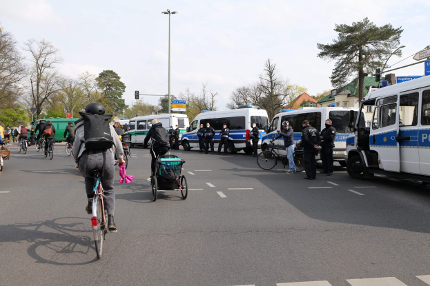 Gruppe von Menschen auf Fahrrädern auf einer Straße mit Polizeiwagen, einige tragen Helme und haben Taschen, andere schieben Kinderwagen, vor einem Hintergrund aus Bäumen, Gebäuden und Verkehrsampeln.