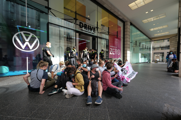 Menschen sitzen vor einem Volkswagen-Autohaus auf dem Boden und halten ein Transparent.