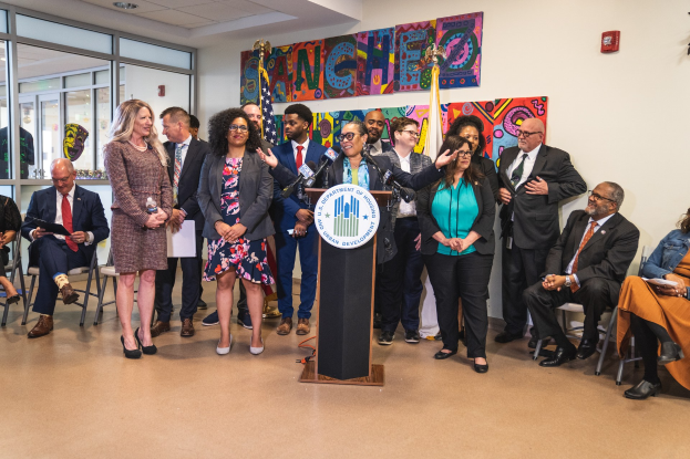 Gruppe von Menschen vor einem Rednerpult mit Mikrofon und einer Tafel mit der Aufschrift "Sancho Stadtrat joins City Council", mit sitzenden und stehenden Teilnehmern, Flaggen, Feuerlöscher und Deckenbeleuchtung im Hintergrund.