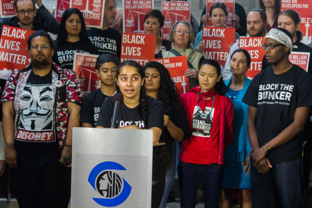 Eine Gruppe von Menschen mit "Black Lives Matter"-Schildern vor einer Menge, mit einer Frau, die auf einem Podium spricht und einer Wand im Hintergrund.