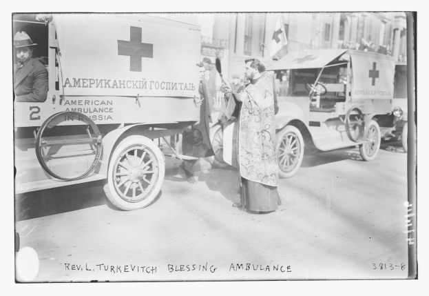 Altes Schwarz-Weiß-Foto eines Mannes neben einem Krankenwagen mit anderen Fahrzeugen und Menschen auf einer Straße, Gebäuden und einer Fahne im Hintergrund und Text unten.