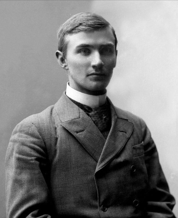 Schwarzes und weißes Foto eines Mannes in Anzug und Krawatte, der mit gefalteten Händen und ernstem Gesichtsausdruck vor der Kamera steht.
