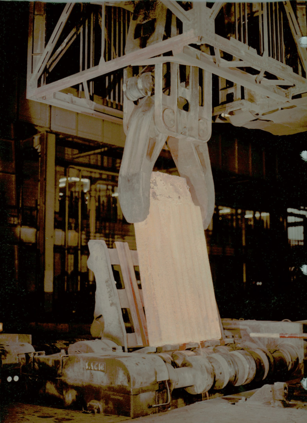 Altes Foto eines großen Krans, der ein Metallförderband in einer Fabrik hochhebt, mit einem Metallobjekt im Vordergrund und einem Gebäude im Hintergrund.