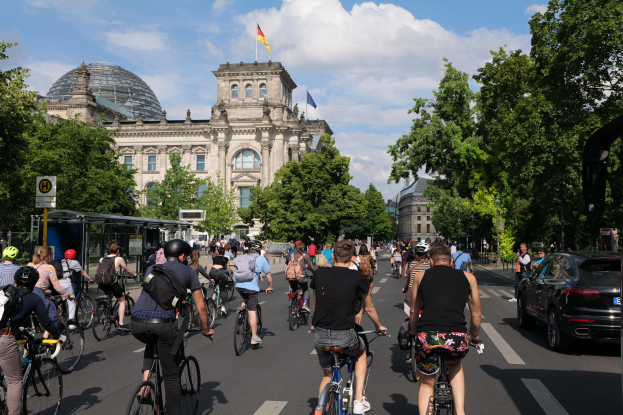Eine Gruppe von Menschen, die auf Fahrrädern eine von Bäumen gesäumte Straße in Berlin, Deutschland, entlangfahren, mit Gebäuden und einer Bushaltestelle, unter einem bewölkten Himmel und einer Flagge auf einem Gebäude.