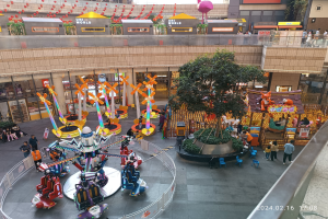 Ein Vergnügungspark innerhalb eines Einkaufszentrums mit verschiedenen Attraktionen, Bäumen, Menschen, Stühlen, Ständen und Gebäuden im Hintergrund, mit einem Wasserzeichen in der rechten unteren Ecke.