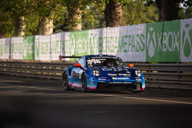 Ein blauer Porsche 911 GTS auf einer Rennstrecke, umgeben von einem Zaun und Bannern, mit Bäumen und einem klaren blauen Himmel im Hintergrund.