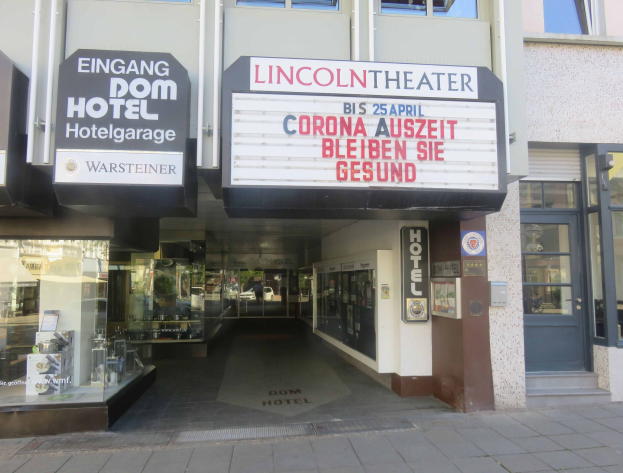 Außenansicht des Lincoln Theaters in Berlin, Deutschland, mit Glasfenstern und -türen sowie einer Texttafel und einer belebten Stadtlandschaft im Inneren.